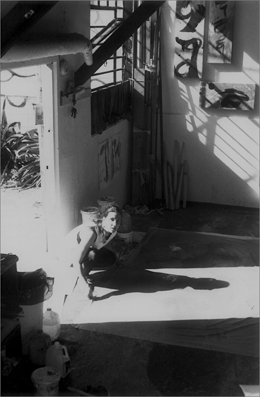artist brenda heim kneeling in the doorway of her studio looking up at the photographer atop a ladder